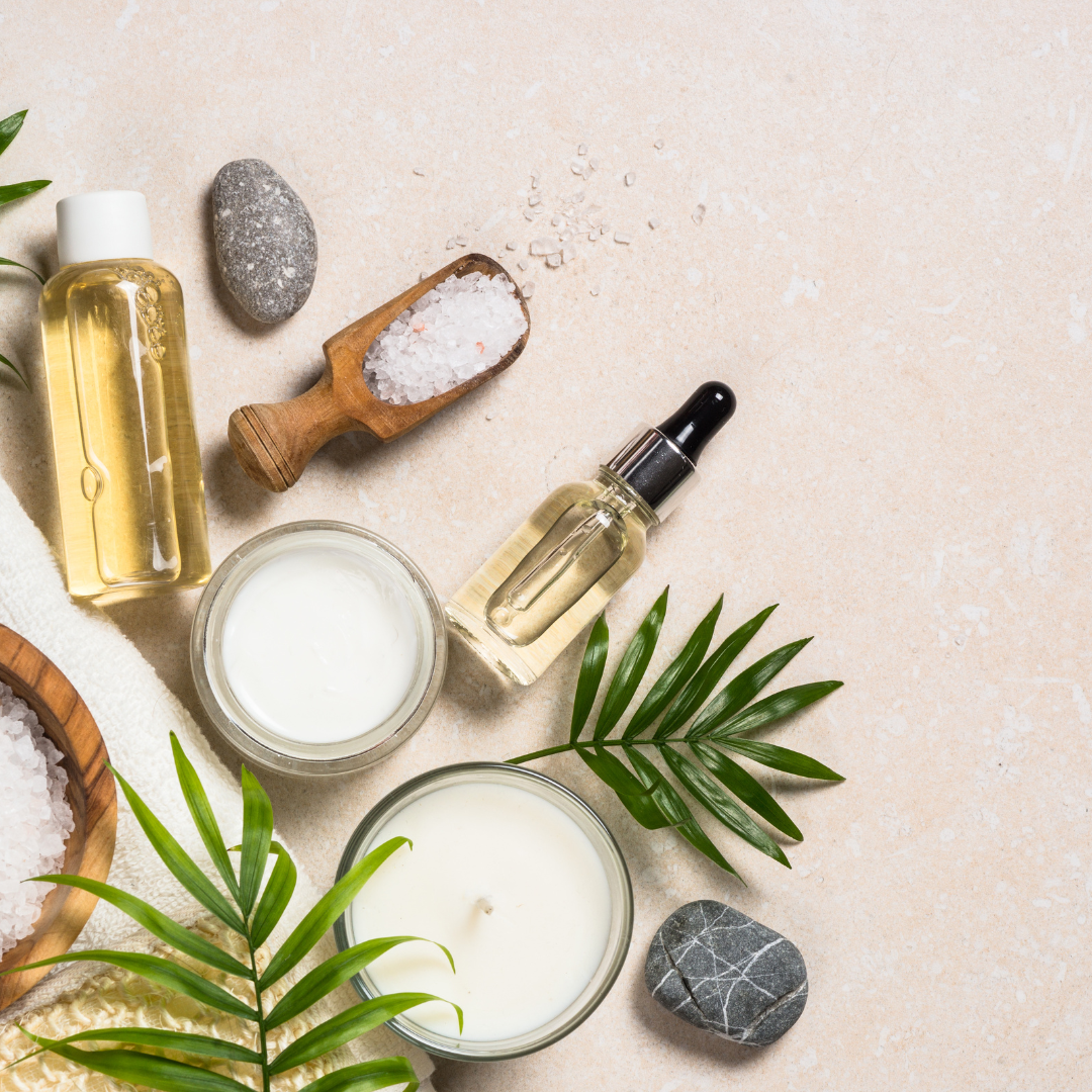 Cosmetic products including a bottle of oil, a jar of cream, a candle, and a dropper bottle on a light surface with green leaves.